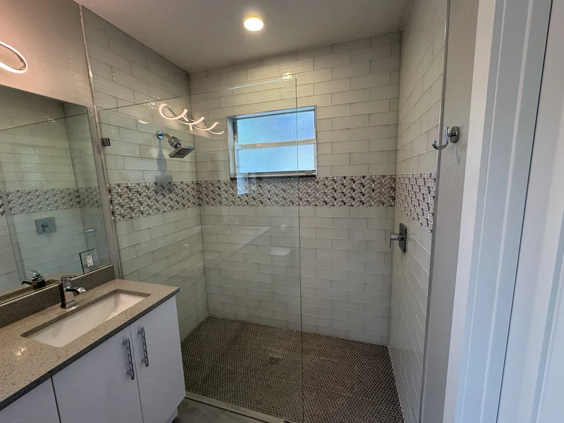 Glass shower beside a double vanity with a patterned tile band and high windows