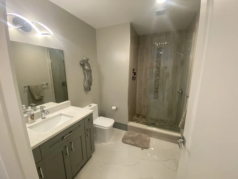 Gray vanity, illuminated mirror and a glass shower with warm textured tile