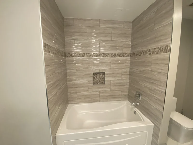 White bathtub surrounded by warm textured wall tile with a mosaic accent strip