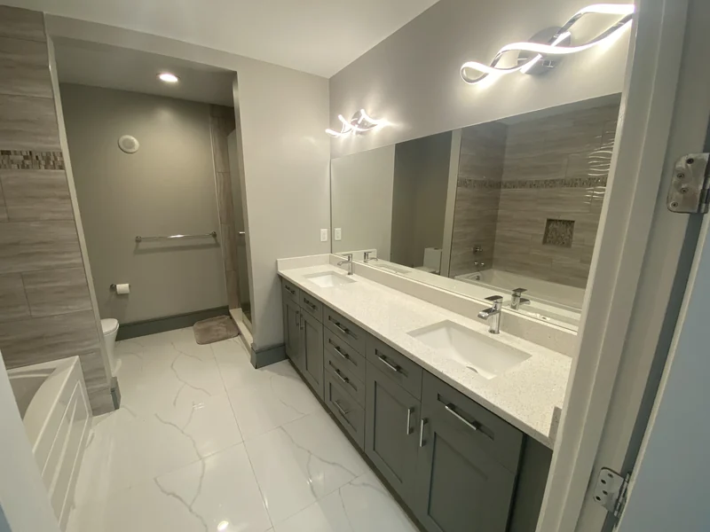 Gray double vanity beside a bathtub and glass shower with warm-toned tile